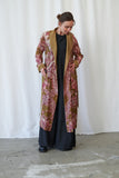 Person wearing a long, patterned coat over a black outfit against a white curtain background