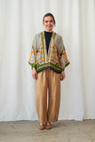 Woman wearing a patterned cardigan and beige pants against a white curtain background