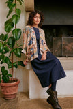 Woman sitting on a bench wearing a patterned jacket and navy dress, with a plant beside her.