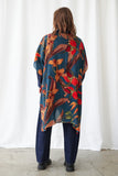 A person viewed from the back wearing a loose, long-sleeve button-up shirt with a bold botanical print in teal, orange, red, and brown over dark blue jeans and black shoes, standing against a white curtain backdrop.